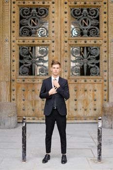 Handsome cauasian groom standing near wooden door and straightening tie, wearing black suit. Concept of male fashion.