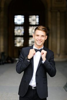 Young european man standing and posing, wearing black suit and bow tie, window in background. Concept of fashionable businessman and male model.