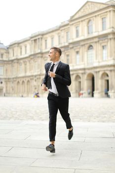 Young good looking man wearing black suit running in Paris, building in background. Concept of business wearstyle, male fashionable model and groom.