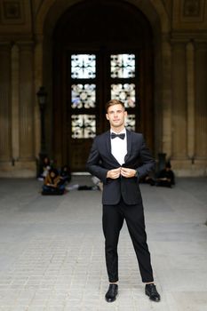 Young european male person standing and posing, wearing black suit and bow tie, window in background. Concept of fashionable businessman and male model.