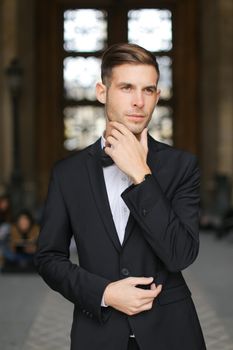 Young caucasian man standing and waiting, wearing black suit and bow tie, window in background. Concept of fashionable businessman and male model.