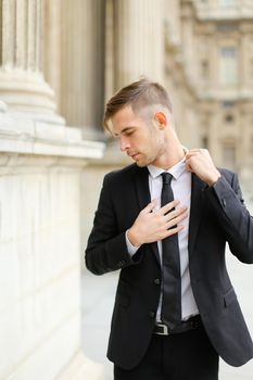 Young handsome man wearing black suit and straightening tie walking in european city, columns in background. Concept of groom fashionable model.