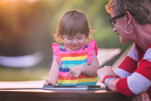 Happy mother and her little daughter enjoying free time using tablet computer while relaxing  on holiday home garden during sunny day