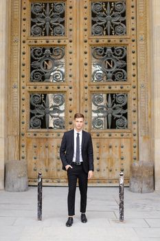 Handsome young caucasian man stnding near wooden door and wearing black suit with tie. Concept of male fashion.