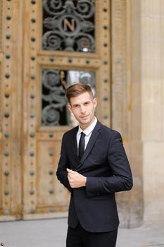 Handsome cauasian groom standing near wooden door and wearing black suit with tie. Concept of male fashion.