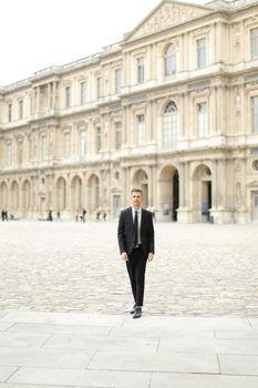 Young good looking man wearing black suit walking in Paris, building in background. Concept of business wearstyle, male fashionable model and groom.
