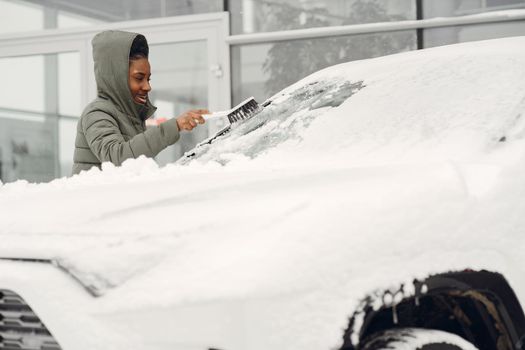 Winter portrait of african woman cleaning snow from a car. Woman in a green jacket.