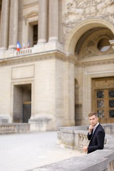 Young european groom waiting and leaning on concrete banister near building. Concept of wedding male photo session or businessperson. Man wearing black suit.