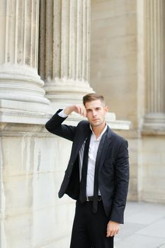 Young caucasian european man wearing black suit with tie leaning on column of building. Concept of groom and male fashionable model.