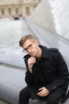 Young man wearing black jacket sitting near glass Louvre Pyramid in Paris, France. Concept of male fashion model and urban photo session.