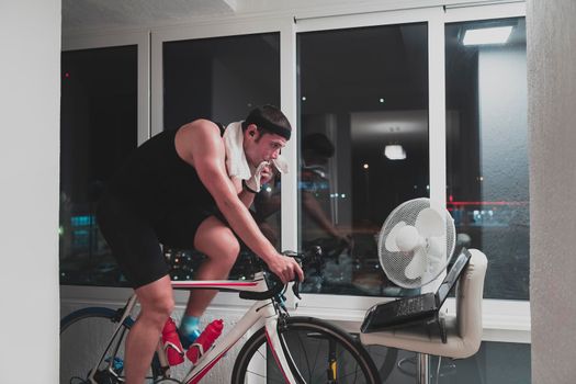 Man cycling on the machine trainer he is exercising in the home at night. Playing online bike racing game during coronavirus covid19 lockdown. New normal concept.