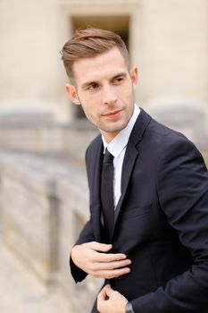 Portrait of young man wearing black suit and leaning on concrete banister. Concept of of waiting groom and male wedding photo session.