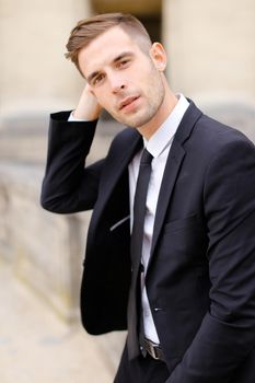 Portrait of handsome man wearing black suit and leaning on concrete banister. Concept of of waiting groom and male wedding photo session.