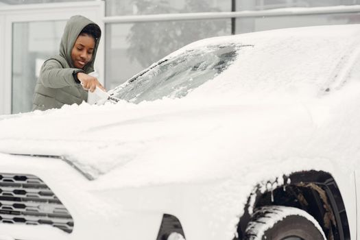 Winter portrait of african woman cleaning snow from a car. Woman in a green jacket.