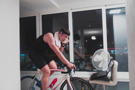 Man cycling on the machine trainer he is exercising in the home at night. Playing online bike racing game during coronavirus covid19 lockdown. New normal concept.