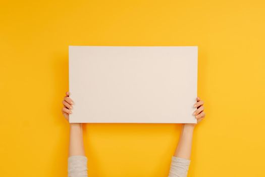 Hands holding white canvas paper blank with copyspace on yellow background. Empty sheet banner with place for text. Concept of design and creativity