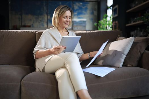 Cheerful middle aged woman sitting on sofa, using computer tablet apps, looking at screen,reading good news in social network, shopping or chatting online.