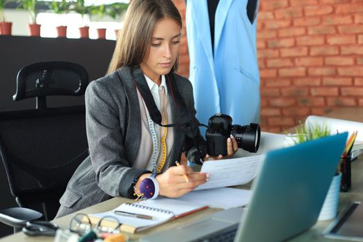 Beautiful young woman looking at her notes and holding digital camera while sitting in her workshop
