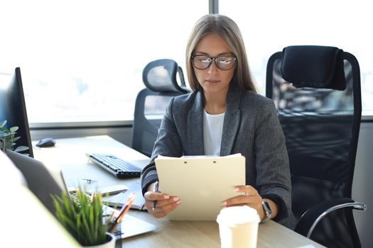 Attractive business woman holding documents and looking at them while sitting at the desk in office.