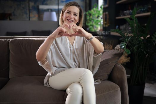 Smiling middle aged woman is showing heart gesture with two hands and looking at camera sitting on sofa