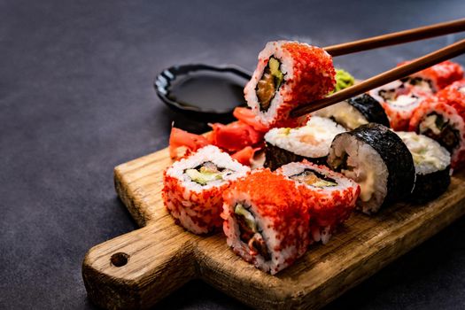 Closeup view of sushi maki set served on wooden tablet. Person hand holding roll with chopstick. Japanese traditional food composition with prepared rice and seafood