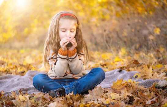 Nice little girl with cup sitting on picnic mat in autumn park