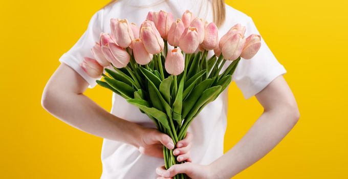 Girl child with pink tulip flowers isolated on yellow background with copyspace. Closeup studio portrait of kid holding bouquet in her hand behind her back