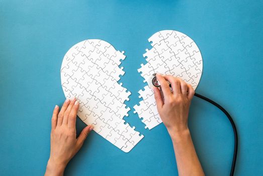 Hands with stethoscope and white puzzle in heart shape. White details of puzzle on blue background. Heart health concept.