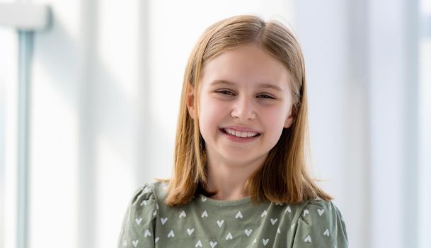 Closeup portrait of preteen beautiful blond hair girl in light room with blurred background. Female kid schoolgirl looking at the camera and smiling indoors