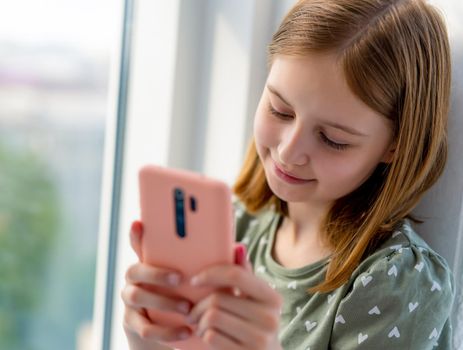 Pretty preteen girl looking at smartphone and chatting with friends at home close to window with daylight. Beautiful kid with cell phone texting indoors