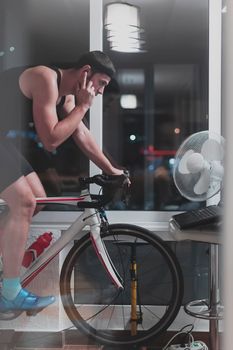 Man cycling on the machine trainer he is exercising in the home at night. Playing online bike racing game during coronavirus covid19 lockdown. New normal concept.