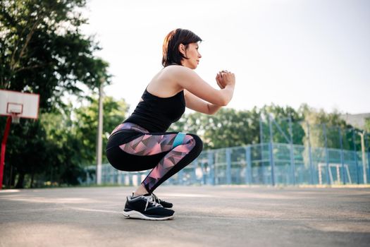 Young girl woman exercising outdoors with mini rubber elastic band doing squats at stadium. Active training for female legs and buttocks with additional sport equipment outside