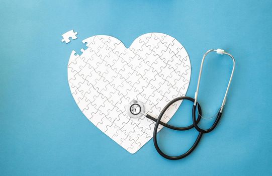 White puzzle in heart shape and stethoscope on blue background. Heart health concept