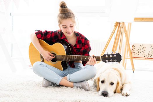 Pretty teenage girl playing guitar next to cute dog at home