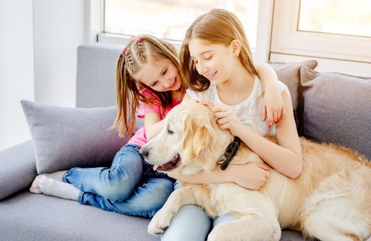 Happy sisters cuddling cute dog sitting on sofa at home