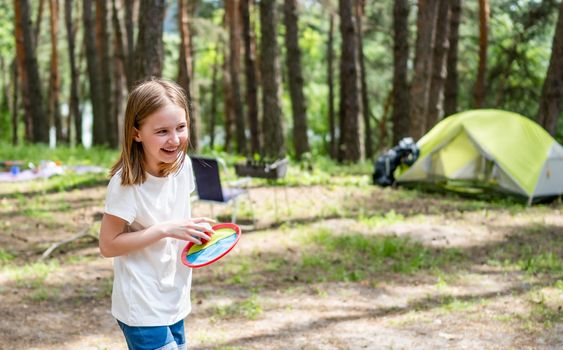 Little girl playing in the camping in the wood with green tent on background. Beautiful smiling kid having activities in the forest with sun light
