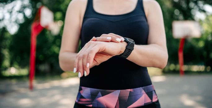 Young girl woman standing at stadium and looking at fitness tracker during workout outdoors. Closeup view of female hands with smart watch for sport training control