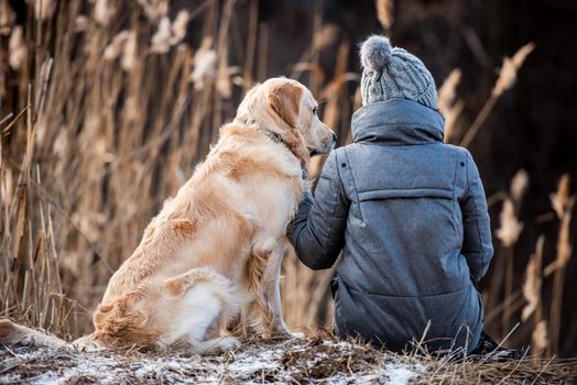Woman owner sitting on the ground with golden retriever dog during early spring walk outdoors . Portrait from the back of girl with doggy pet labrador together at the nature