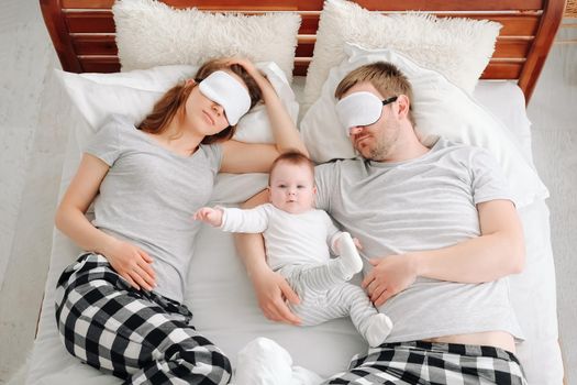 Family with child lying in the bed together wearinf same pajamas and eye masks and sleeping. Young mother and father hugging toddler kid and resting in the bedroom