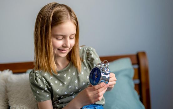 Portrait of preteen beautiful girl looking at metal clock with blue clockface in bedroom. Female kid schoolgirl sitting in the bed and turn on alarm