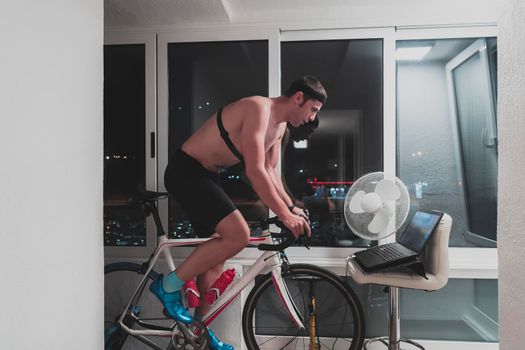 Man cycling on the machine trainer he is exercising in the home at night. Playing online bike racing game during coronavirus covid19 lockdown. New normal concept.