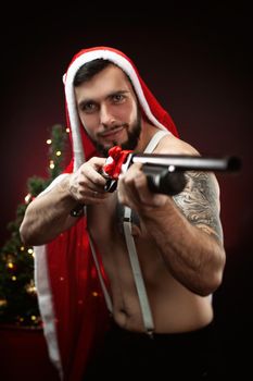 strong guy in a red Santa Claus cape at the Christmas tree with a shotgun in his hands