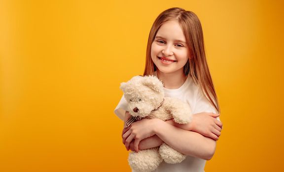 Beautiful little girl child hugging teddy bear and smiling isolated on yellow background with copyspace. Horizontal portrait of kid holding toy and looking at the camera