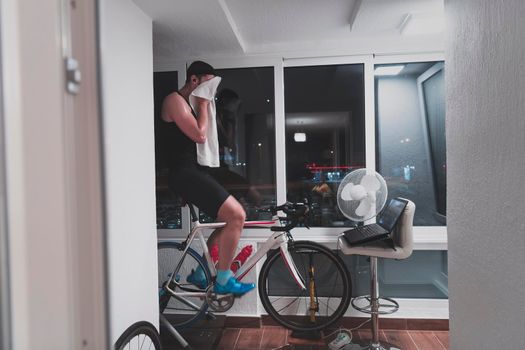 Man cycling on the machine trainer he is exercising in the home at night. Playing online bike racing game during coronavirus covid19 lockdown. New normal concept.