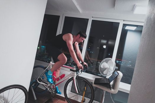 Man cycling on the machine trainer he is exercising in the home at night. Playing online bike racing game during coronavirus covid19 lockdown. New normal concept.