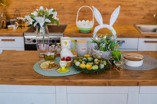 Still life of painted Easter eggs in wreath with lemon and olives and tomatoes on wooden kitchen counter. A lovely basket with bunny ears with flowers and decorative eggs. Vase of lilies on the counter.