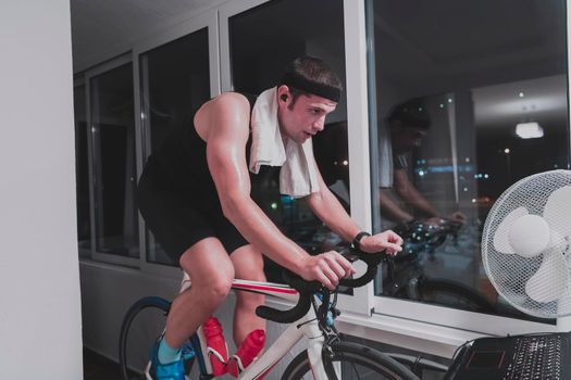 Man cycling on the machine trainer he is exercising in the home at night. Playing online bike racing game during coronavirus covid19 lockdown. New normal concept.