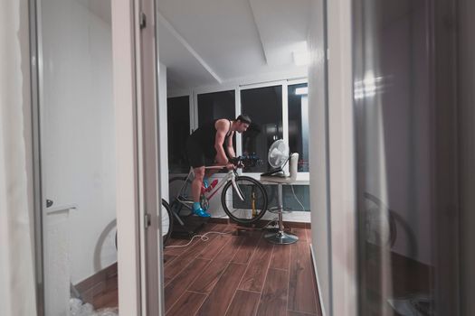 Man cycling on the machine trainer he is exercising in the home at night. Playing online bike racing game during coronavirus covid19 lockdown. New normal concept.