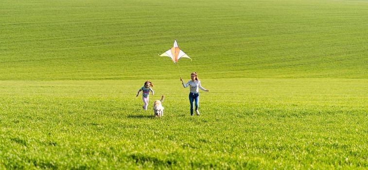 Two girls and dog running on the green field and holding flying kite