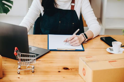 Entrepreneur owner using pen point note and laptop checking online purchase shopping order to preparing pack product box. Packing goods for delivery to customer